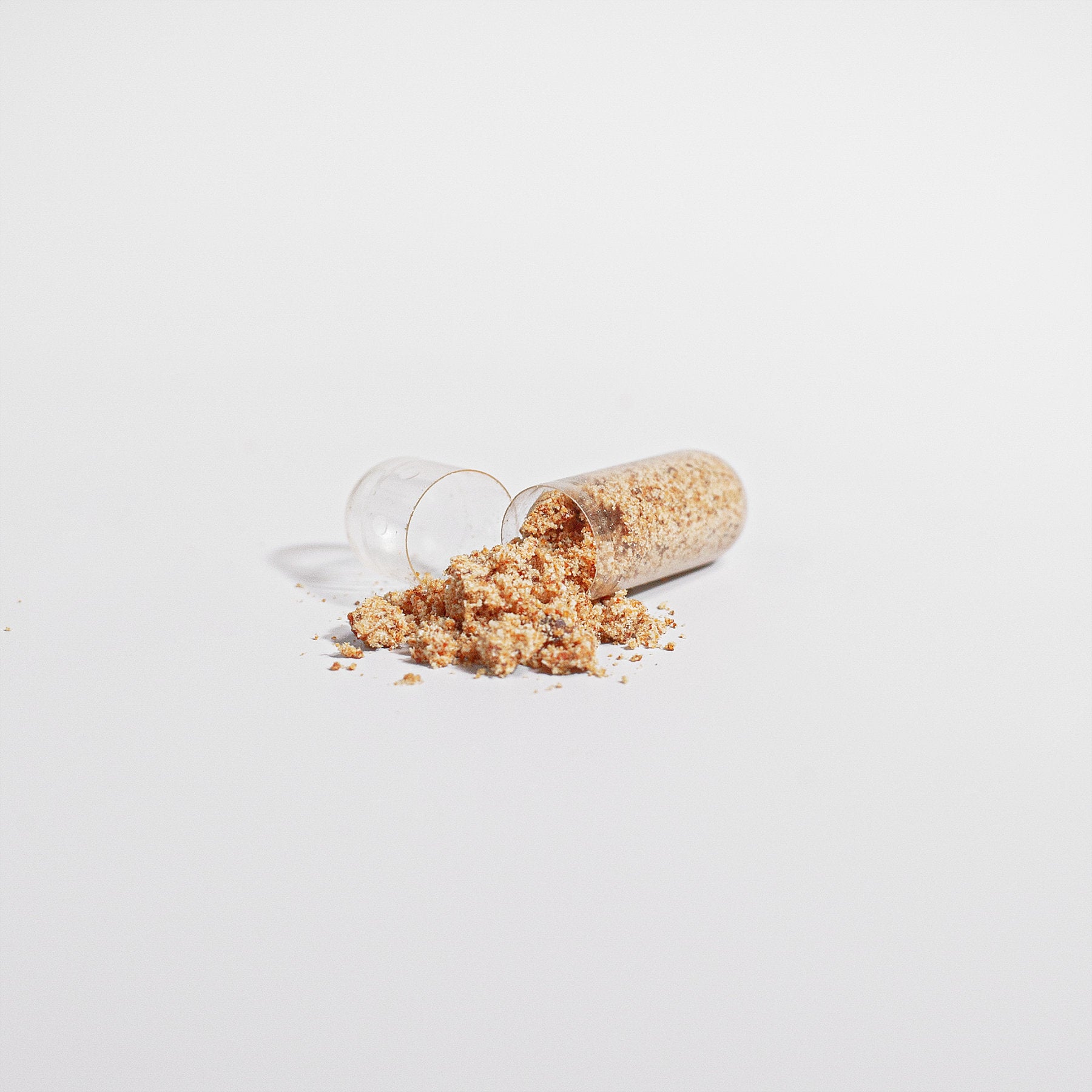 A small glass vial is tipped over, causing a gluten-free granular substance to spill onto a white surface. The tan and beige material scatters in a small pile near the vial&