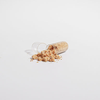 A small glass vial is tipped over, causing a gluten-free granular substance to spill onto a white surface. The tan and beige material scatters in a small pile near the vial&