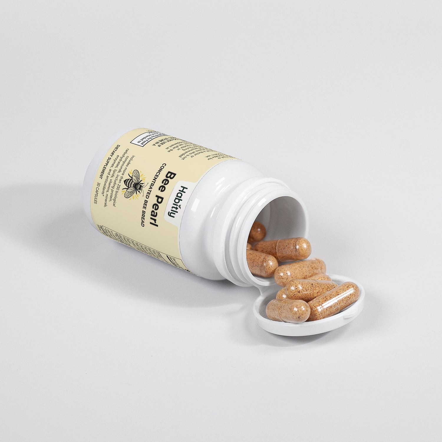 A beige bottle labeled &quot;Bee Pearl Concentrated Bee Bread Capsules&quot; by Habitly lies on its side with the lid off, revealing several tan capsules spilling onto the white surface. The label, featuring a bee image, indicates that the contents are gluten-free supplements.