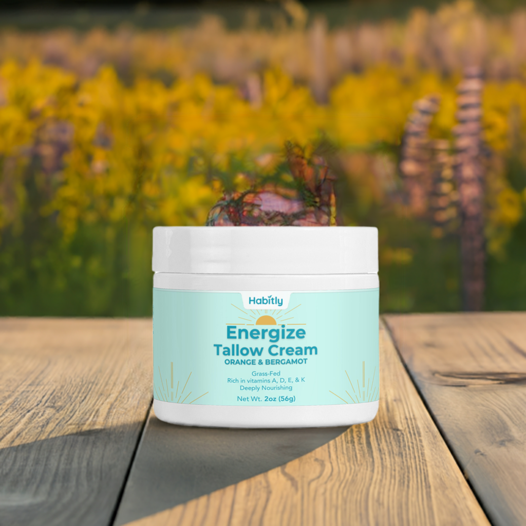 A jar of Habitly Energize Tallow Cream with Orange &amp; Bergamot, crafted using grass-fed beef tallow, rests on a wooden surface. The label emphasizes its natural skincare benefits and the invigorating scent of orange and bergamot, enhanced with vitamins A, D, E, and K. Blurry yellow flowers and greenery in the background provide a natural touch.