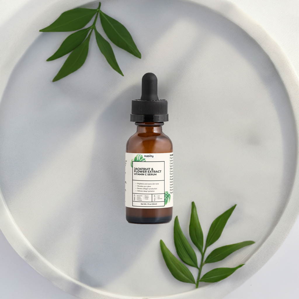 A brown dropper bottle labeled &quot;Jackfruit &amp; Flower Extract Vitamin C Serum&quot; by Habitly rests on a white circular tray, offering antioxidant protection. Green leaves are artistically placed around the bottle, creating a natural and organic look. The minimalist background highlights the product&