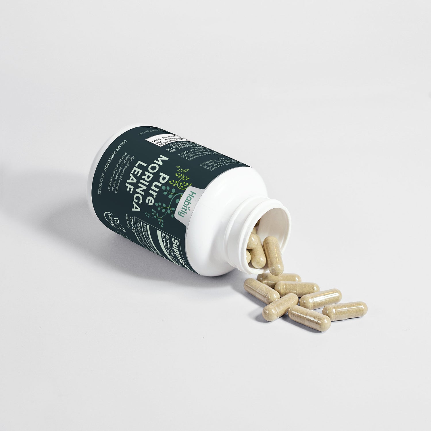 A white bottle branded as &quot;Habitly Pure Moringa Leaf Capsules&quot; with green text is tipped over, spilling beige capsules onto a white surface. The label emphasizes its role as a dietary supplement rich in antioxidants.