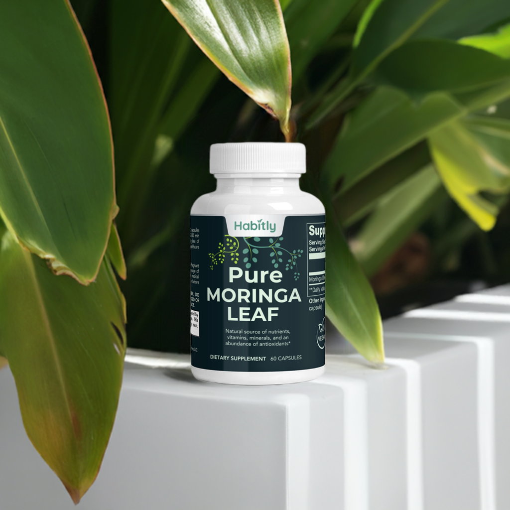 A bottle of Habitly Pure Moringa Leaf Capsules, containing 60 capsules, rests on a white surface surrounded by vibrant green plant leaves. The label highlights its natural content of antioxidants, vitamins, and amino acids.