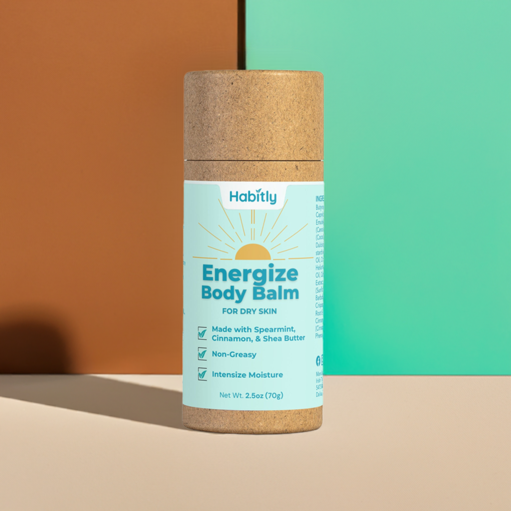 A cylindrical cardboard container of the Habitly Energize Body Balm is displayed against a two-tone brown and teal background. The eco-friendly packaging highlights its non-greasy formula, which is enriched with spearmint, cinnamon, and shea butter to deeply moisturize dry skin.