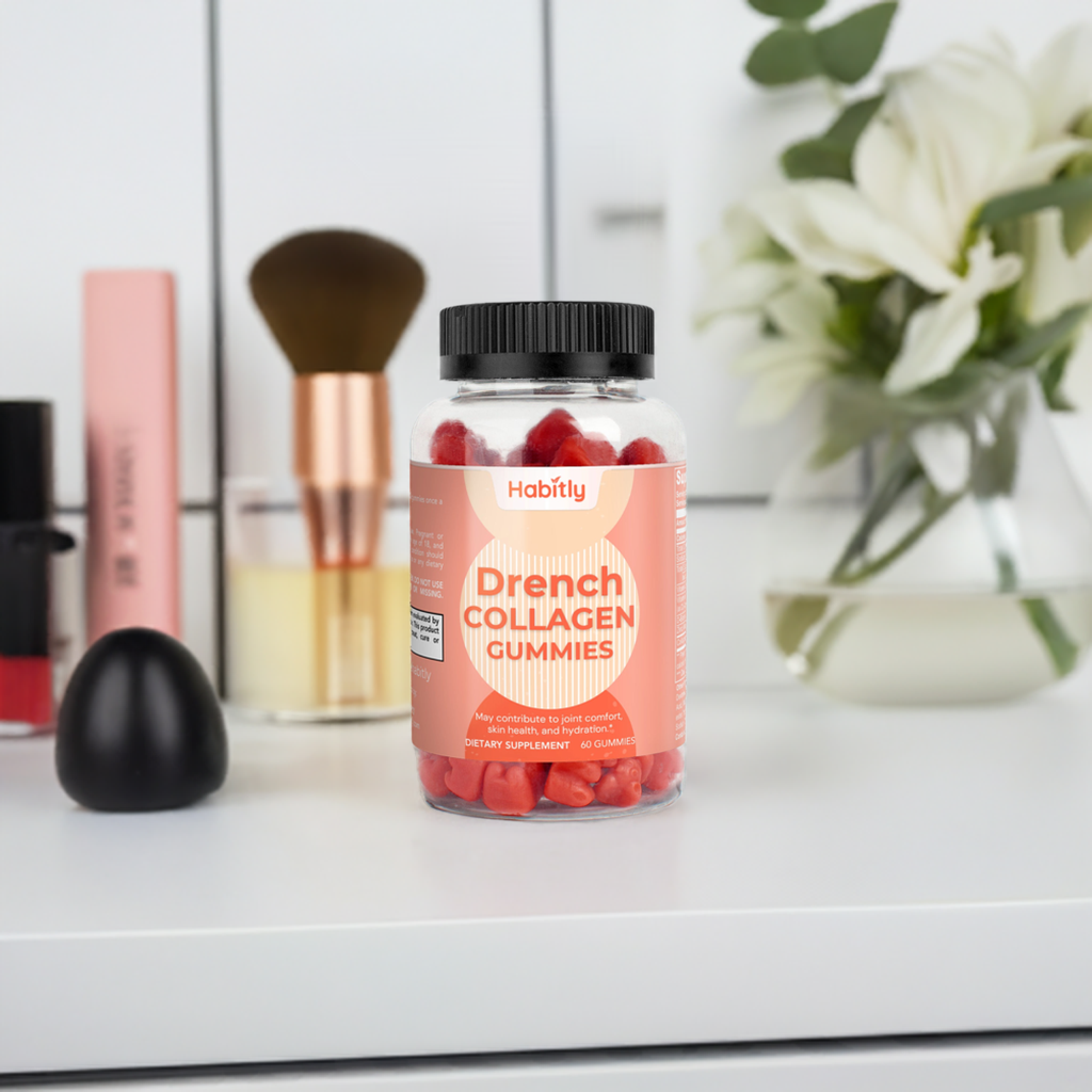 A bottle of &quot;Drench Collagen Gummies&quot; by Habitly is placed on a white surface, offering a promise of Vitamin C enhancement. In the background, makeup items, a brush, and a vase with white flowers set an inviting scene for this dietary supplement.