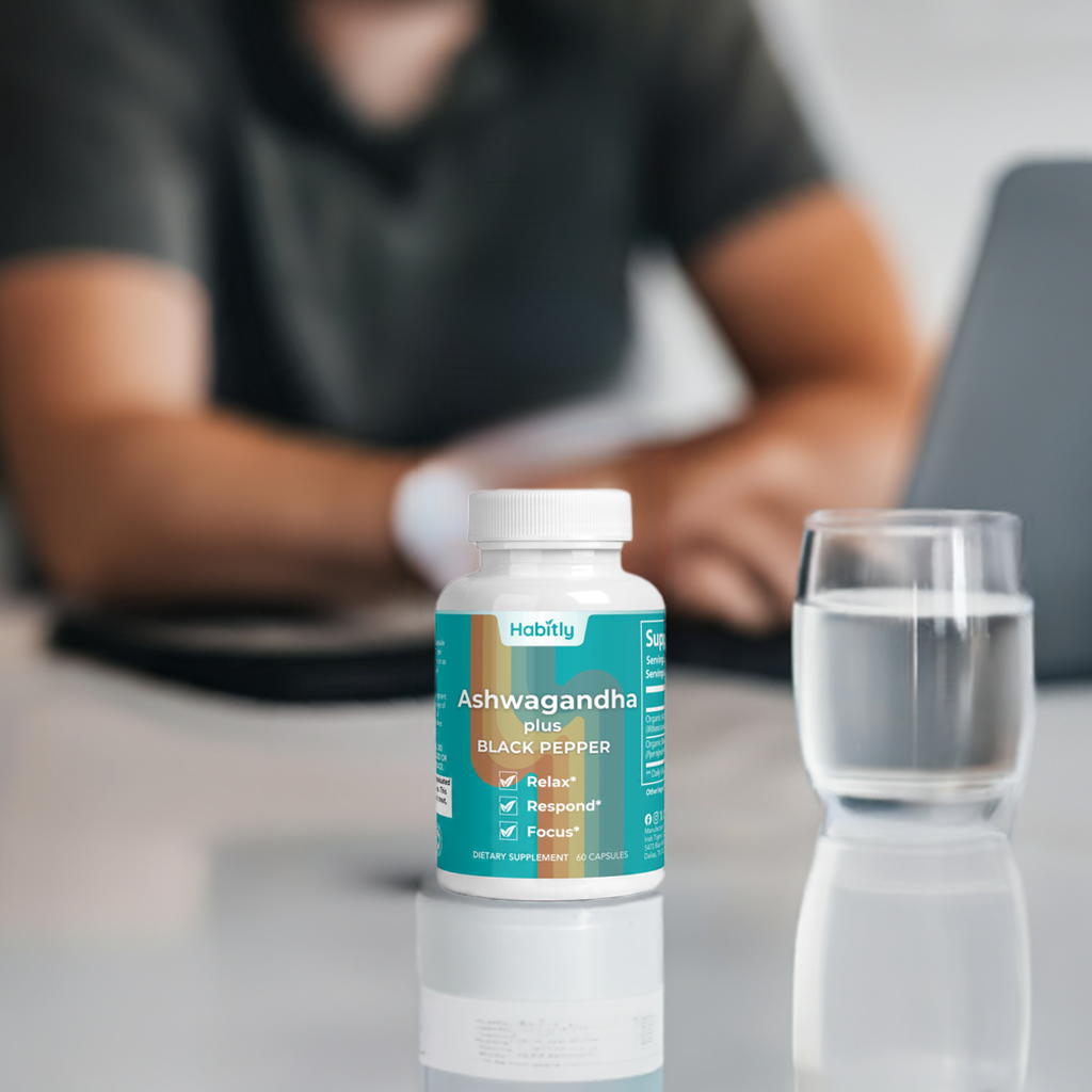 A bottle of Habitly Ashwagandha with Black Pepper, known for its adaptogenic properties, rests on a desk next to a glass of water. In the soft-focus background, someone wearing a gray shirt is busy working on their laptop. The label emphasizes benefits such as relaxation, enhanced response, and improved focus.